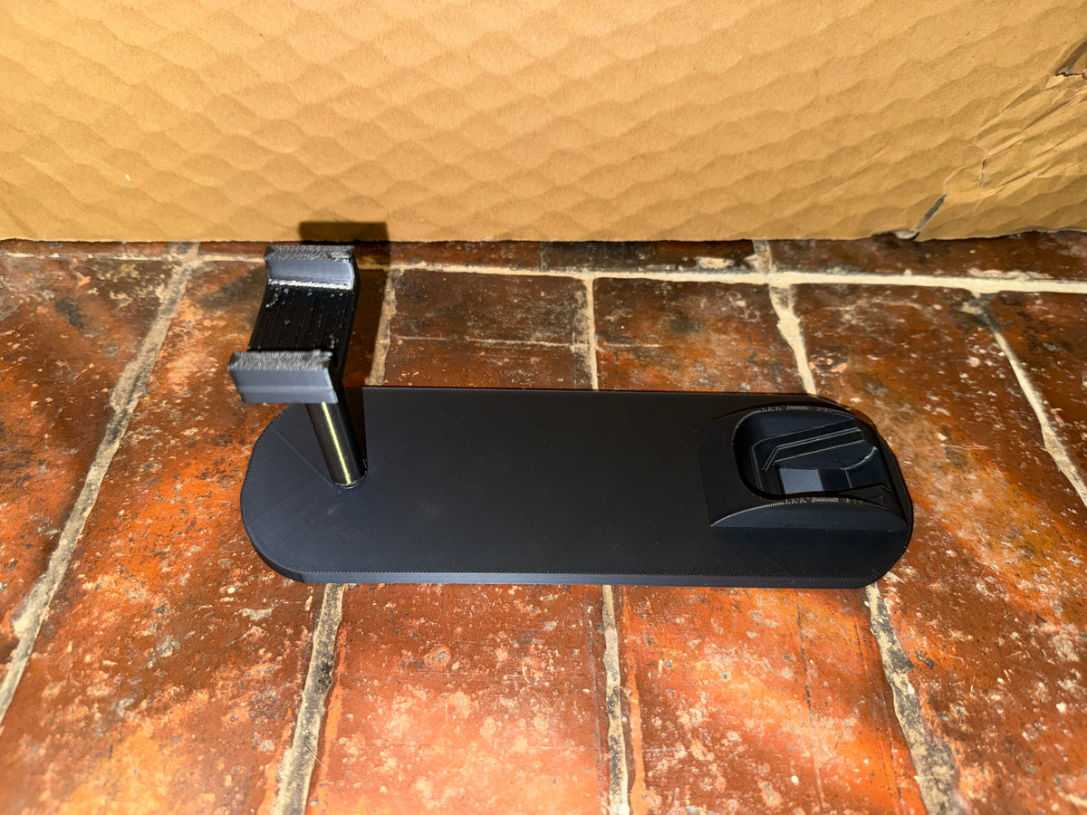 A black rectangular mounting base or stand with a vertical metal rod and bracket, placed on a reddish-brown tiled floor against a tan cardboard wall.