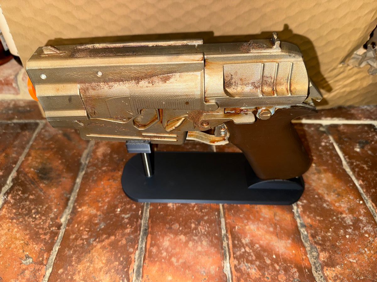 A miniature replica of a sci-fi blaster pistol, styled with a weathered, rusted metallic finish, displayed on a black stand on a brick floor.