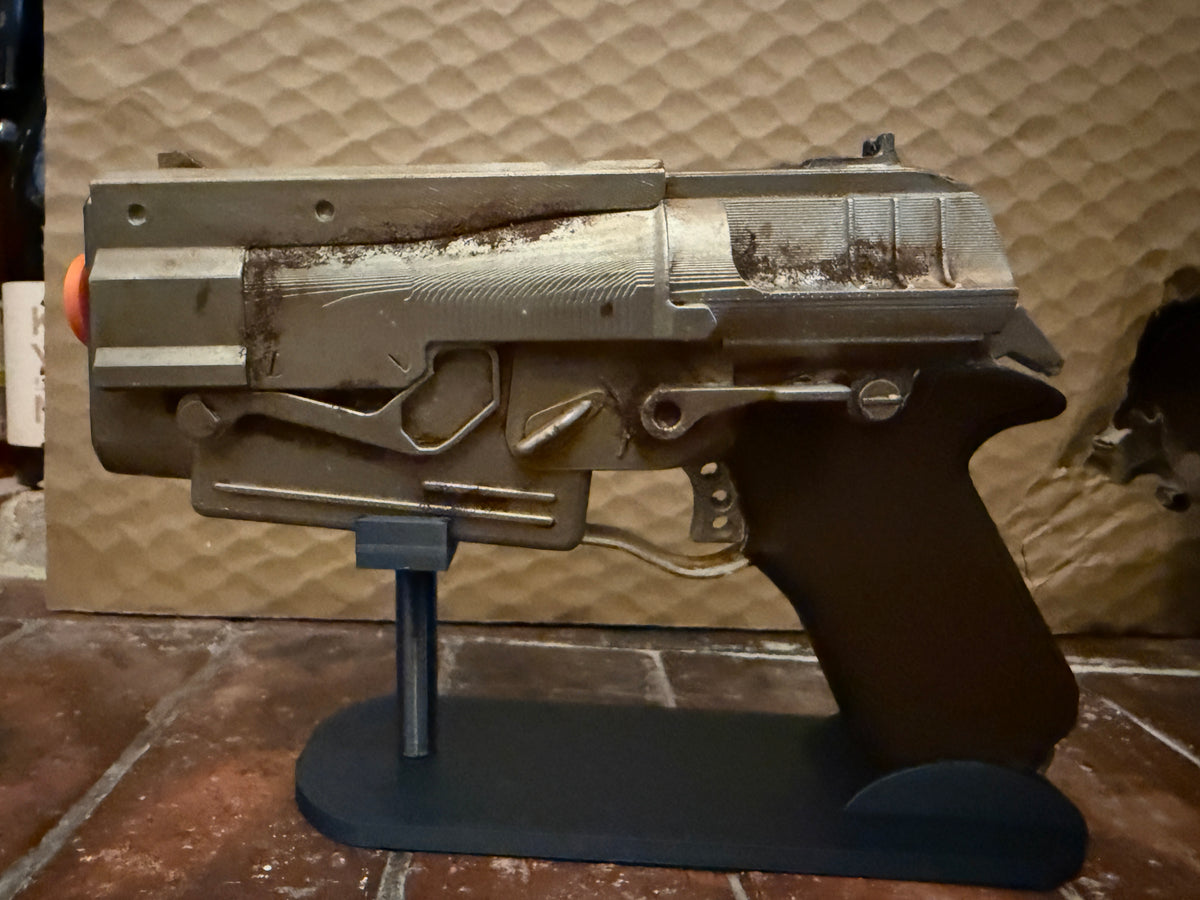 A replica toy blaster pistol with a weathered metallic finish and an orange tip, displayed on a black stand against a textured tan background.
