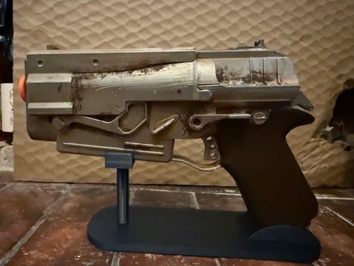 A replica toy blaster pistol with a weathered metallic finish and an orange tip, displayed on a black stand against a textured tan background.