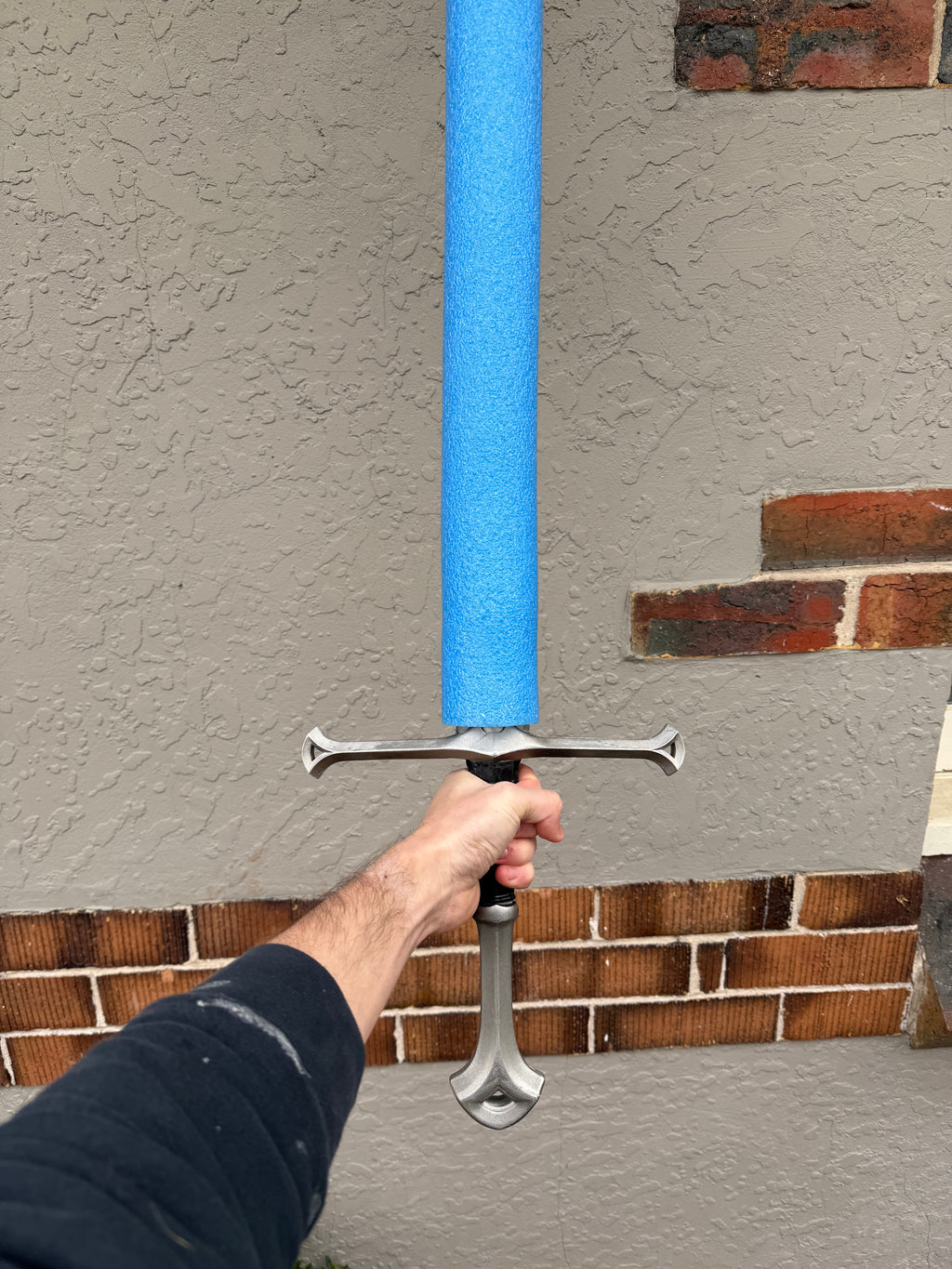 A person's hand holding a foam sword with a bright blue foam blade, a silver crossguard, and a silver hilt, positioned in front of a stucco wall with brick trim.