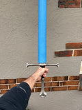 A person's hand holding a foam sword with a bright blue foam blade, a silver crossguard, and a silver hilt, positioned in front of a stucco wall with brick trim.