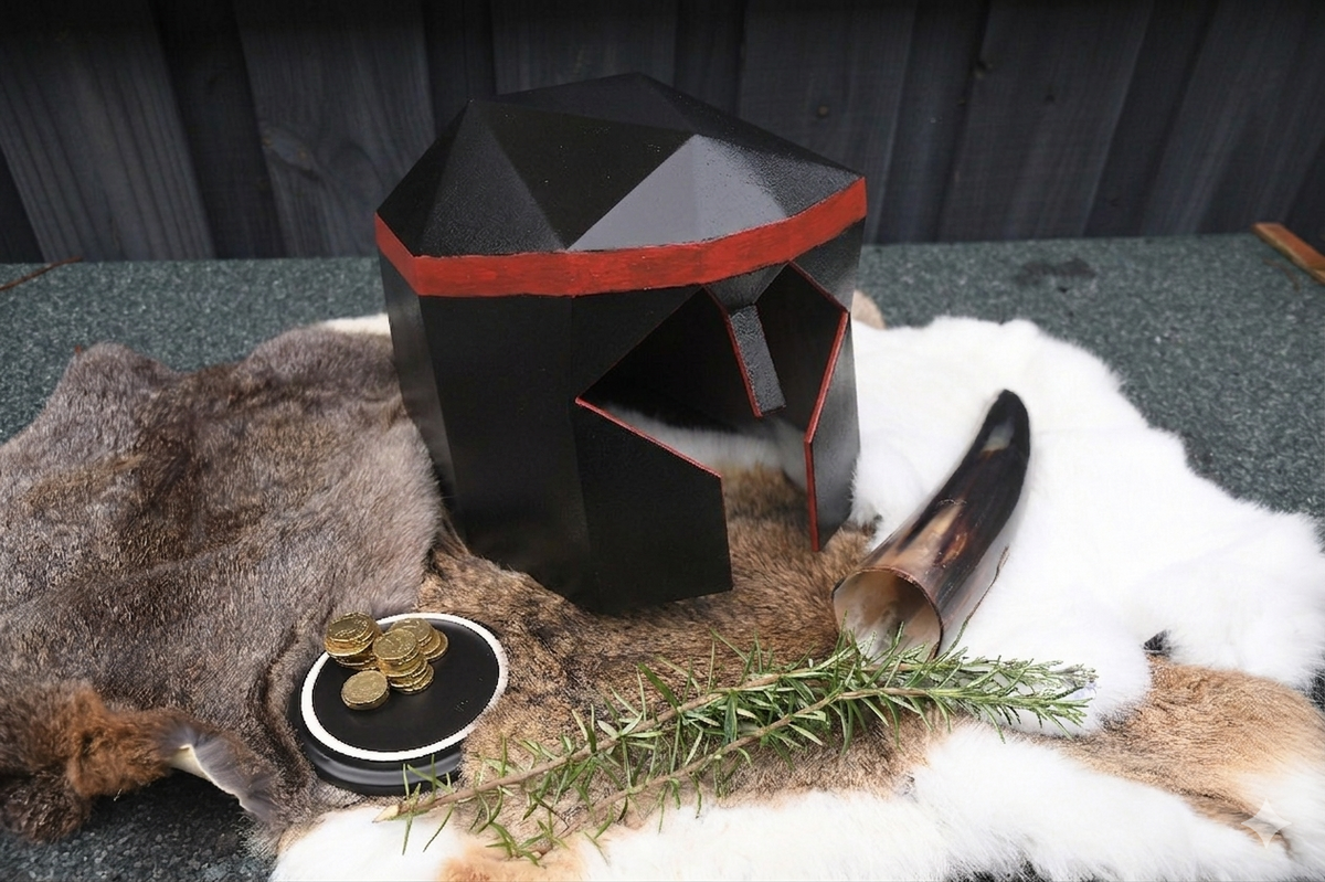 A black geometric helmet-shaped candle holder with red trim, styled on a fur rug with a horn, coins, rosemary, and a small dish on a dark tabletop.